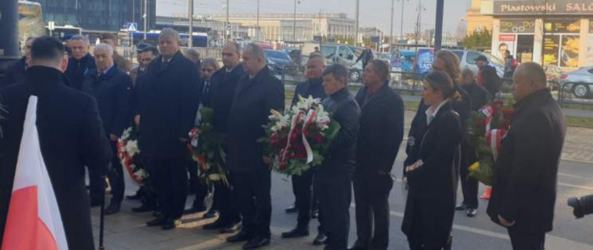 Kilka osób stojących w rzędzie, większość w czarnych, ciemnych płaszczach, w środku osoba trzymająca wiązankę z biało-czerwoną szarfą. Wszyscy stoją na chodniku twarzą do osoby robiącej zdjęcie.