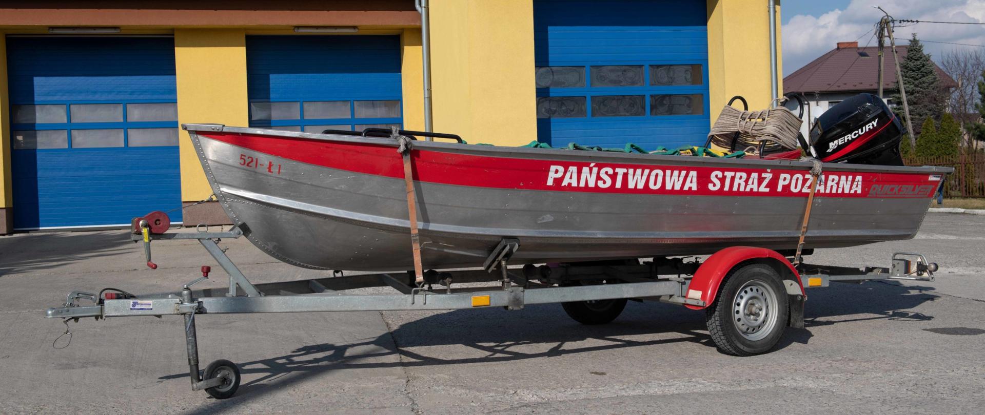 Płaskodenna łódź aluminiowa na przyczepie, lawecie jednoosiowej z zamontowanym silnikiem na tle bram wjazdowych do garażu od strony placu wewnętrznego. Wewnątrz łodzi widoczne nosze lodowe i liny ratownicze.