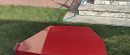 Na zdjęciu widać czerwony zasobnik od pieca leżący na trawniku przed domem. Trawa zielona. Zdjęcie wykonane w ciągu dnia. Schody przed drzwiami wejściowymi.