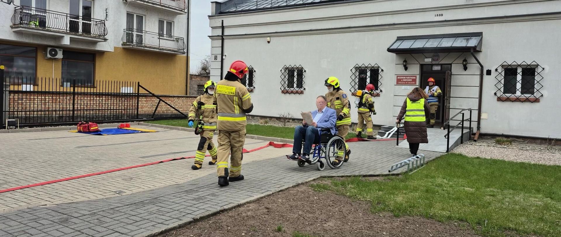 Strażacy pomagają ewakuować starszego mężczyznę na wózku inwalidzkim z budynku; obok stoi osoba w kamizelce odblaskowej, a w tle widoczny jest namiot ratowniczy i sprzęt.