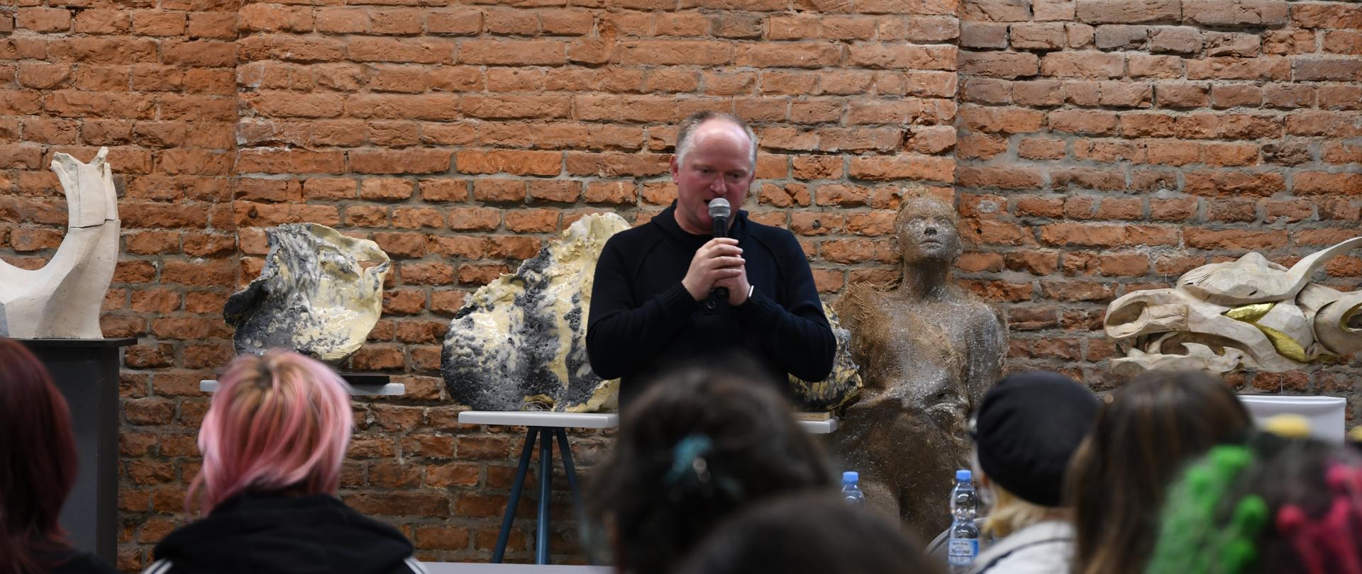 Mężczyzna przemawiający do mikrofonu przed publicznością, w tle rzeźby i ceglana ściana