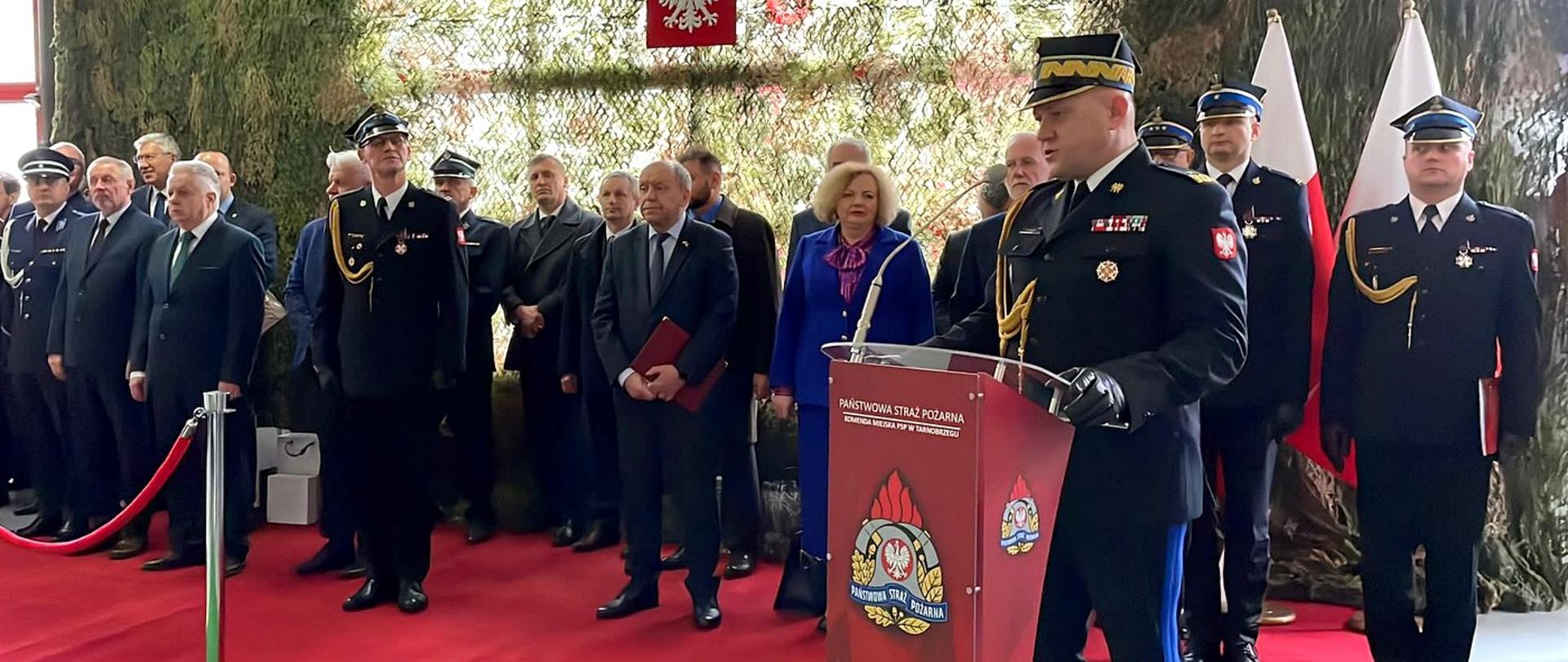 Zdjęcie zrobione w pomieszczeniu podczas uroczystej zbiórki. Przed mównicą stoi funkcjonariusz Państwowej Straży Pożarnej w generała i przemawia. Za nim stoją zaproszeni goście.