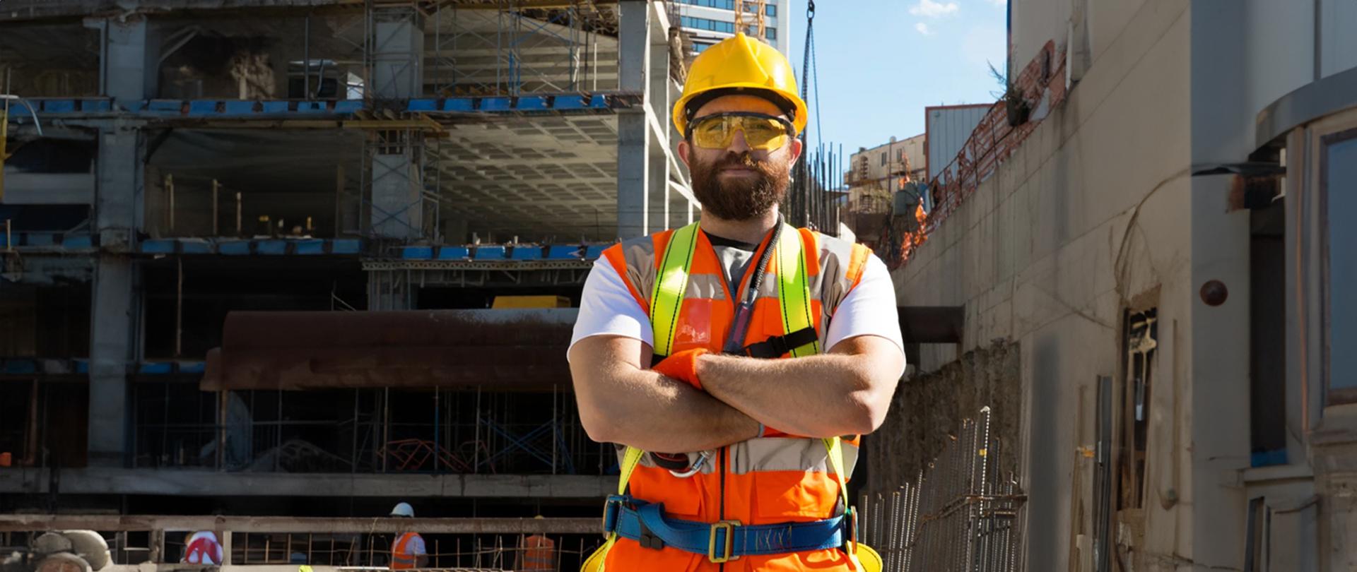 Na zdjęciu pracownik budowy w kasku, kamizelce odblaskowej i okularach ochronnych, który stoi na tle placu budowy. 