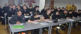 Świetlica KP PSP w Rawiczu. Za pięcioma rzędami stołów zasiadają słuchacze - uczestnicy szkolenia podstawowego OSP. W iekszośc z nich mana sobie koszulki w kolorze czarnym lub tego samego koloru ubrania koszarowee. Wśród słuchaczy sa zarówno kobiety jak i mężczyźni. Rozwiązują test pisemny, mając przed sobą arkusze egzaminacyjne. W tle ściana z logo PSP i nazwą komendy. Z prawej strony dwóch członków komisji egzaminacyjnej w mundurach piaskowych.
