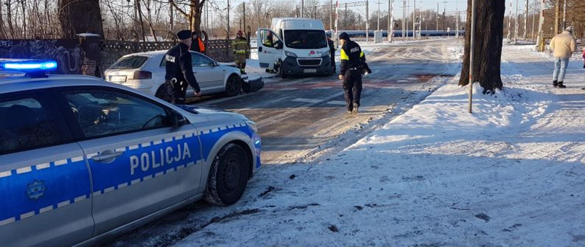 Zdjęcie pokazujące całe miejsce zdarzenia. Wykonane w zimowy dzień. Jest dużo śniegu, droga jest oblodzona. Pierwszy plan stanowi radiowóz policyjny, srebrny z niebieskim paskiem policja. W oddali pojazdy uczestniczące w kolizji. Plan zamyka przejazd kolejowy. Na drodze stoją policjanci a przy busie strażak