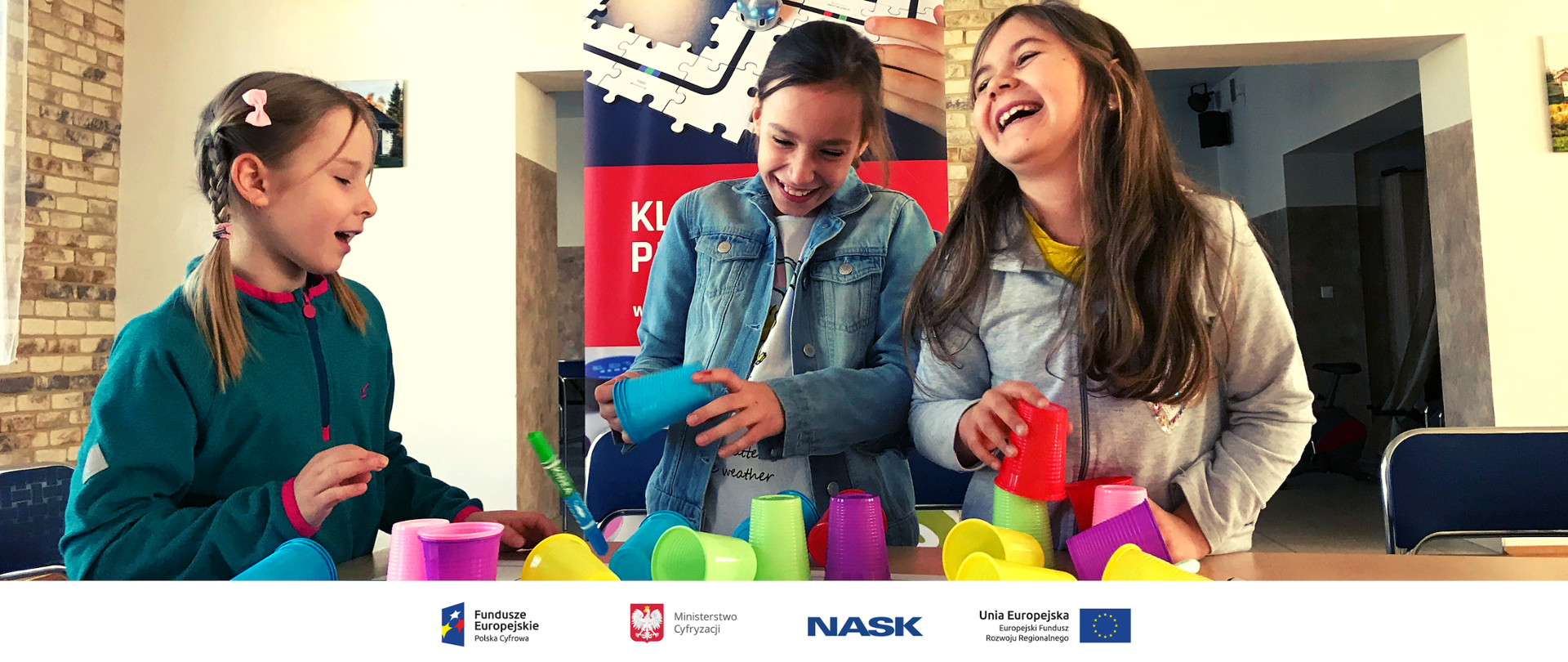 Na zdjęciu widać trzy dziewczynki bawiące się kolorowymi kubeczkami. Na dole zdjęcia umieszczone są logotypy: Fundusze Europejskie. Polska Cyfrowa, Ministerstwo Cyfryzacji, NASK oraz Unia Europejska. Europejski Fundusz Rozwoju Regionalnego. 