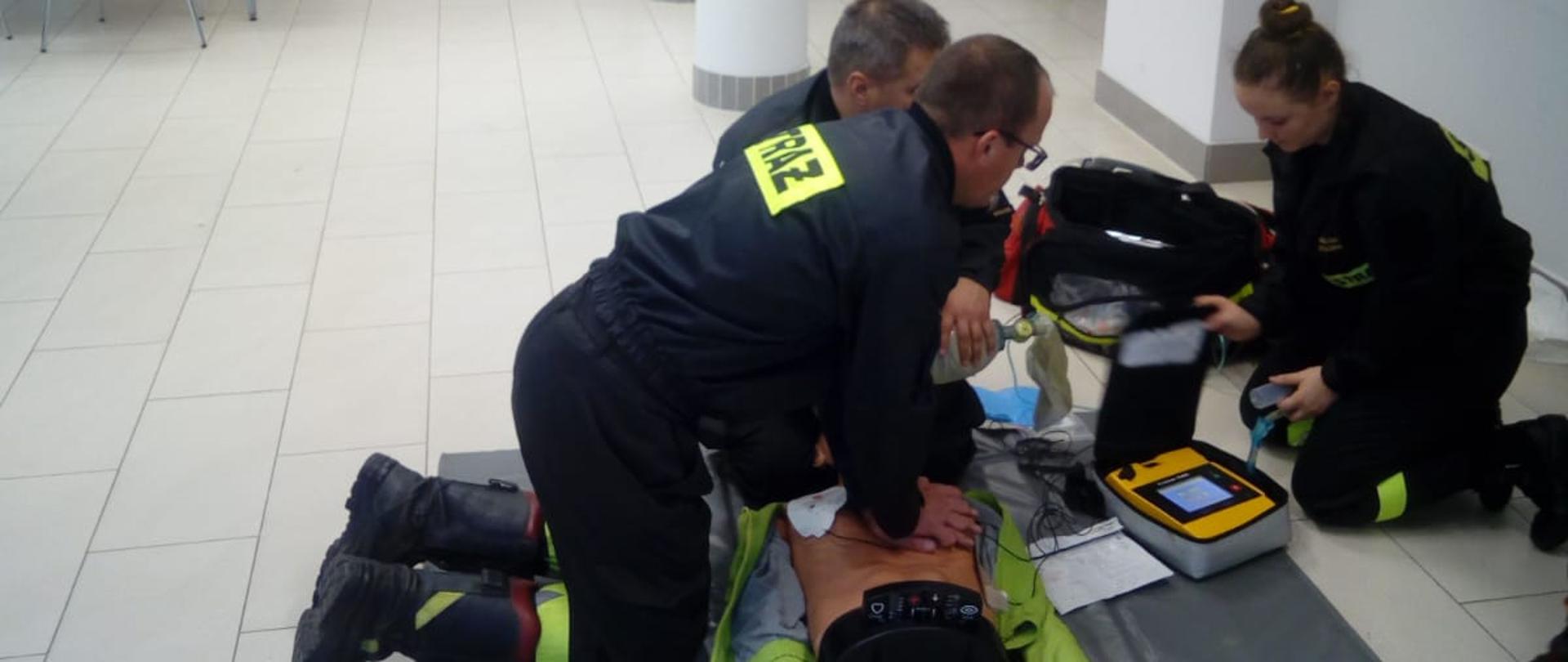 Dwóch mężczyzn i jedna kobieta ubranych w czarny mundur strażacki koszary koloru czarnego tj,: koszule oraz kurtki, klęczą nad manekinem w na Sali konferencyjnej. Mężczyźni i kobieta udzielają kwalifikowanej pierwszej pomocy manekinowi osoby dorosłej podczas egzaminu. W tle na ścianie koloru białego znajduje się sztandar w gablocie oraz żaluzje pionowe koloru szarego
