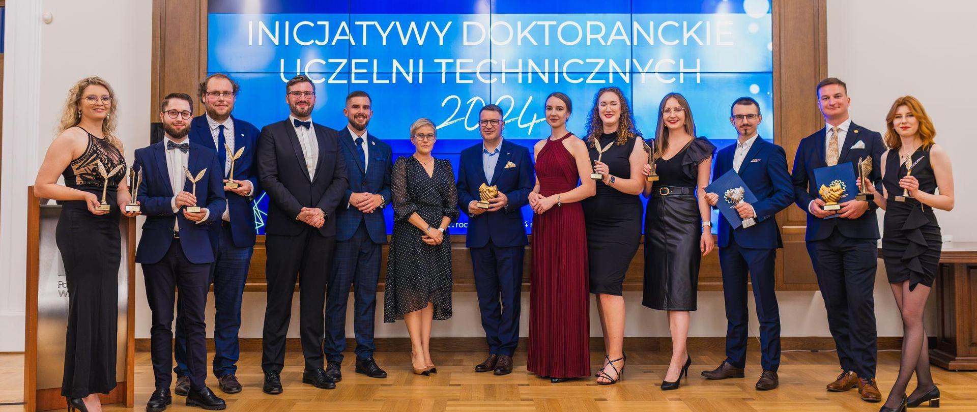 Zdjęcie grupowe, pod wielkim wbudowanym w ścianę ekranem z napisem Inicjatywy doktoranckie uczelni technicznych stoi kilkanaście osób, pośrodku grupy wiceminister Mrówczyńska.