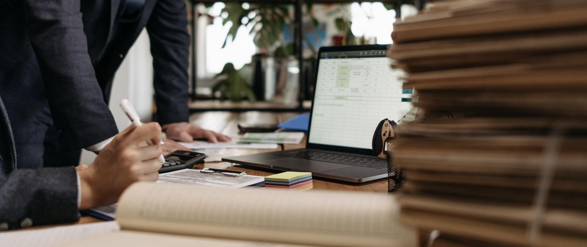 Widok na biurko, stos dokumentów, kalkulator i laptop, w tle dwie osoby - widać ich dłonie, coś notują (źródło: Pexels.com)