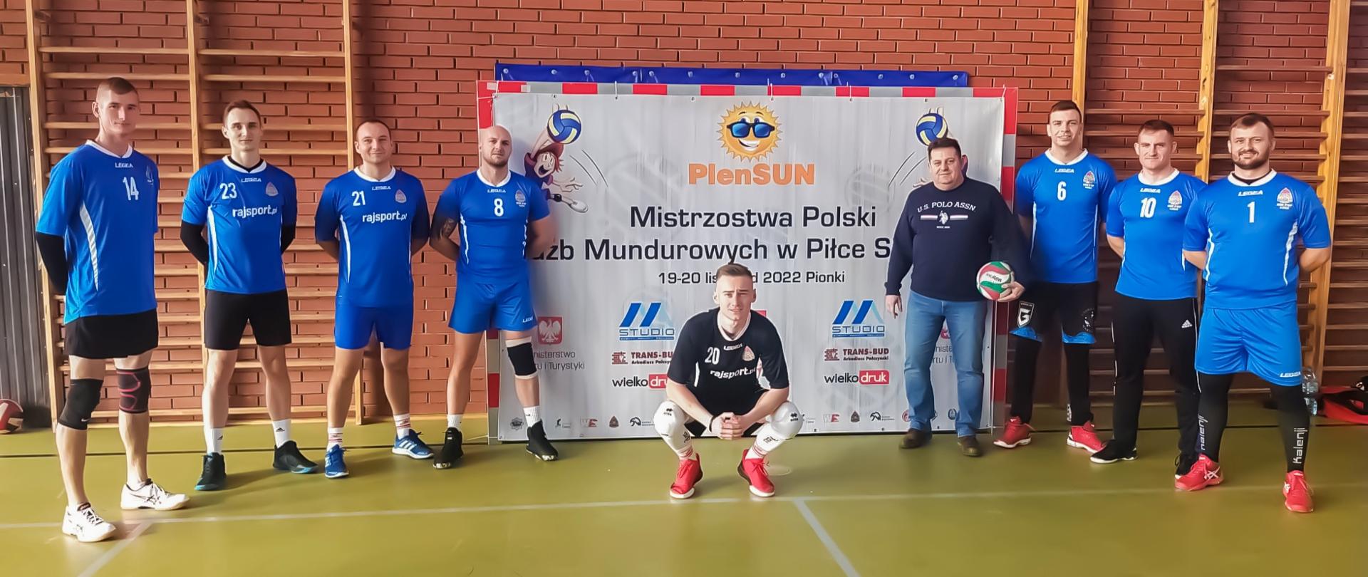 ośmiu siatkarzy oraz kierownik drużyny na tle ceglanej ściany, na ścianie znajdują się: drabinki gimnastyczne oraz baner Mistrzostwa Służb Mundurowych w Piłce Siatkowej