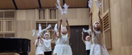 Na scenie stoją uczennice w białych strojach. W uniesionych do góry rękach trzymają białe ozdobne dekoracje przypominające pierzaste skrzydła.