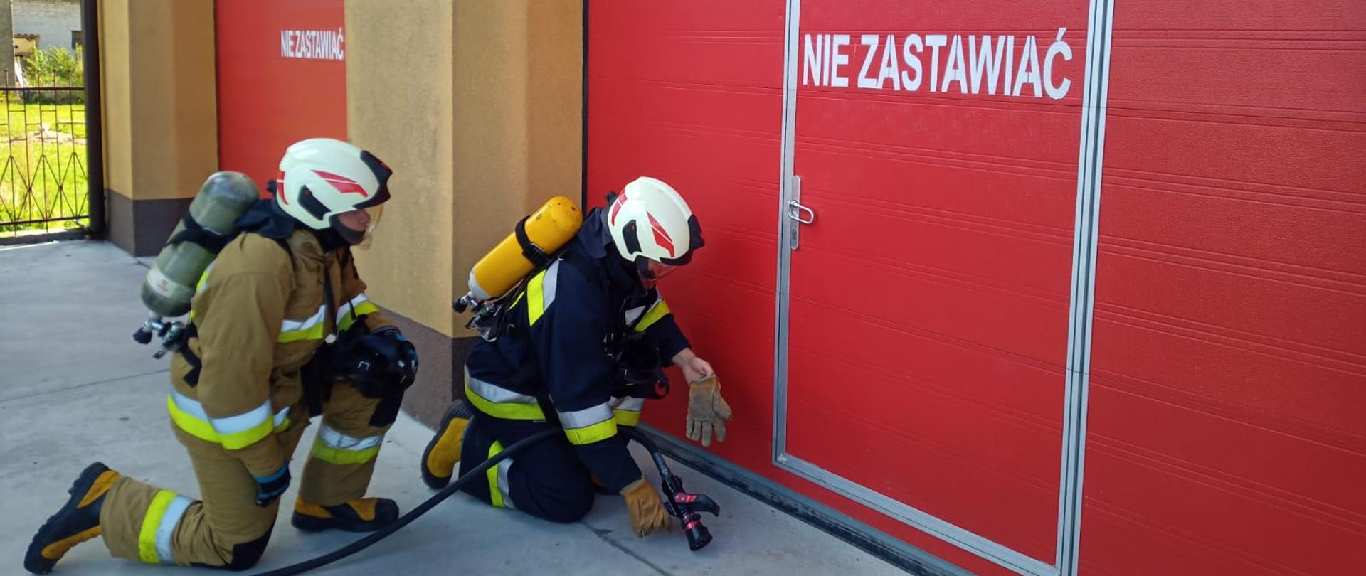 Inspekcje Gotowości Operacyjnej Jednostek OSP KSRG - podczas inspekcji 2 strażacy w ubraniu specjalnym i aparatach ODO przenoszą wąż pożarniczy, patrzą na czerwona bramę garażu.