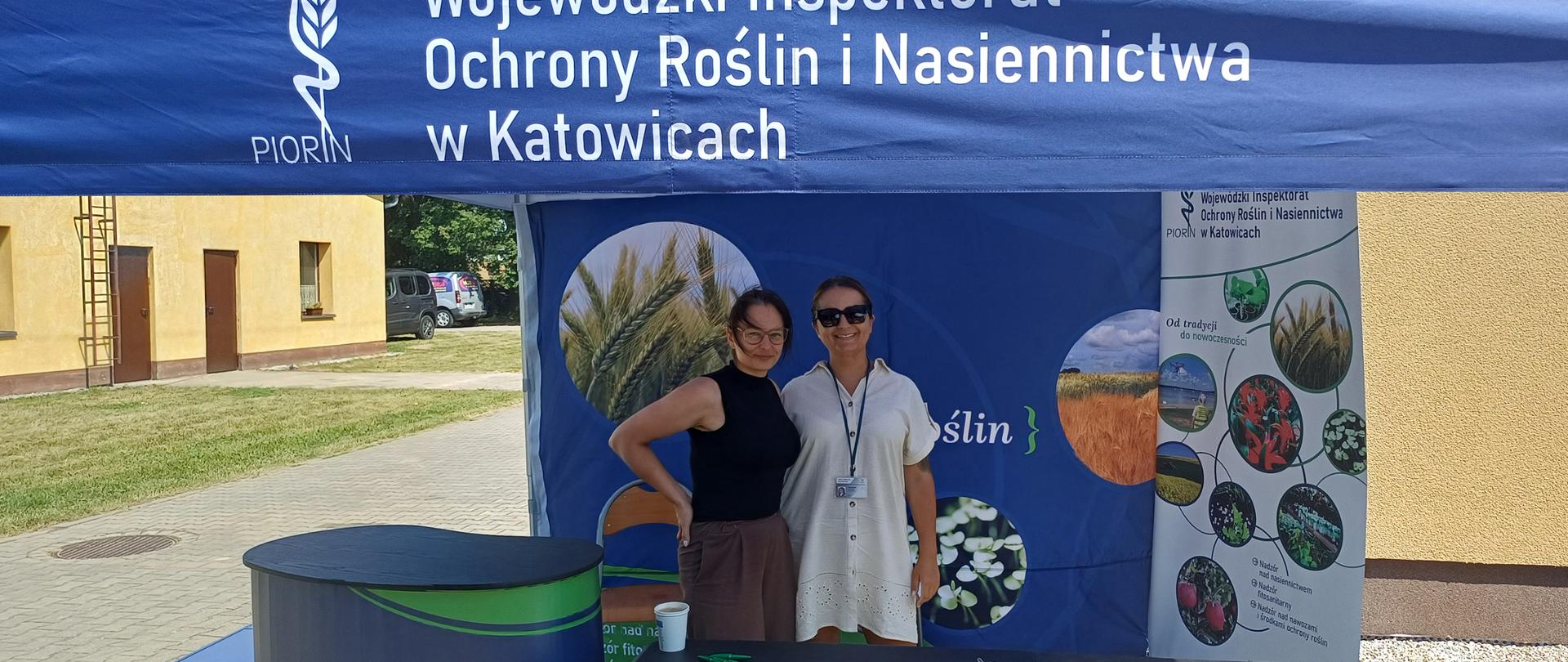  Na zdjęciu widać stoisko promocyjno-informacyjne Wojewódzkiego Inspektoratu Ochrony Roślin i Nasiennictwa w Katowicach, zorganizowane pod dużym, granatowym namiotem. Na froncie namiotu znajduje się duży biały napis: „Wojewódzki Inspektorat Ochrony Roślin i Nasiennictwa w Katowicach” oraz logo PIORiN przedstawiające stylizowany kłos i laskę Eskulapa.
>
> W centralnej części kadru, za stołem wystawowym, stoją dwie uśmiechnięte kobiety pozujące do zdjęcia. Osoba po lewej ma ciemne włosy związane z tyłu, okulary oraz ubrana jest w czarny, bezrękawnikowy golf i ciemne spodnie. Jedną rękę opiera na biodrze. Osoba po prawej ma ciemne okulary przeciwsłoneczne, jasną, krótką sukienkę lub długą koszulę z kołnierzykiem oraz identyfikator zawieszony na szyi.
>
> Przed nimi znajduje się długi, ciemny stół, na którym rozłożono broszury, ulotki, foldery informacyjne i długopisy. Po lewej stronie stoi okrągły, granatowy kontuar z logo PIORiN. Na stole widoczny jest także biały kubek jednorazowy.
>
> Tło stoiska tworzy niebieska ścianka z fotografiami roślin uprawnych i pól oraz fragmentem napisu odnoszącego się do ochrony roślin. Po prawej stronie ustawiono roll-up z dodatkowymi zdjęciami upraw, szkodników i roślin oraz informacjami o działalności Inspektoratu.
>
> W tle, poza namiotem, widoczny jest jasny budynek z kilkoma drzwiami i oknami, fragment parkingu z samochodem oraz teren pokryty trawą i kostką brukową. Zdjęcie wykonano w słoneczny dzień.
