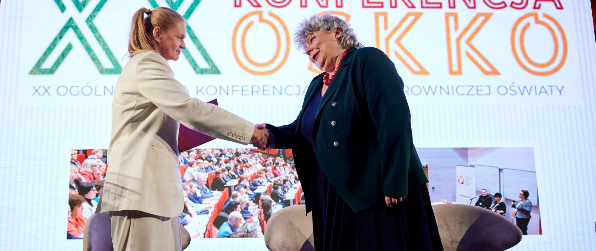 Minister Barbara Nowacka stoi na scenie, podaje rękę nowej małopolskiej kurator oświaty. W tle telebim z napisem XX Ogólnopolska Konferencja Kadry Kierowniczej Oświaty 