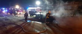 Na zdjęciu strażacy gaszą samochód zadymienie za nimi inni strażacy i samochody ratowniczo - gaśnicze 