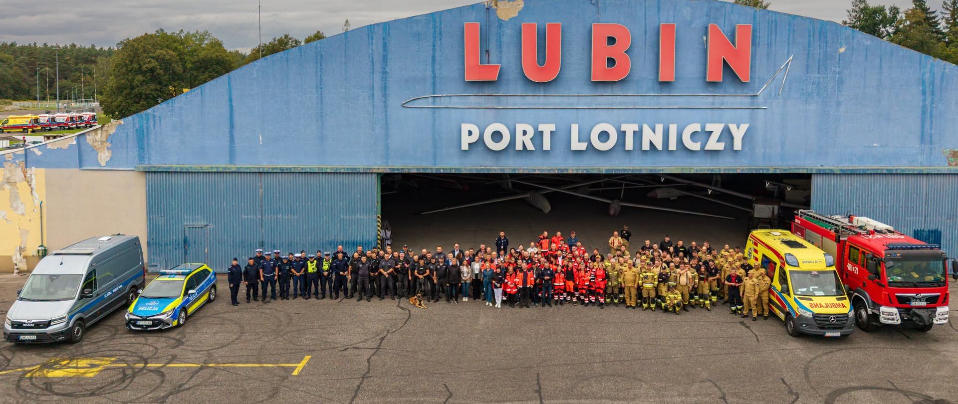 Zdjęcie z drona przedstawiające dużą grupę przedstawicieli służb ratunkowych i mundurowych pozujących przed niebieskim hangarem z napisem „LUBIN PORT LOTNICZY”. Po obu stronach grupy zaparkowane są pojazdy służbowe: radiowozy policji, ambulans oraz czerwony wóz strażacki.