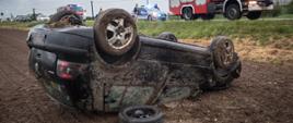 Samochód osobowy marki Volkswagen koloru czarnego leży w polu na dachu. W tle samochód straży pożarnej oraz radiowóz policyjny.