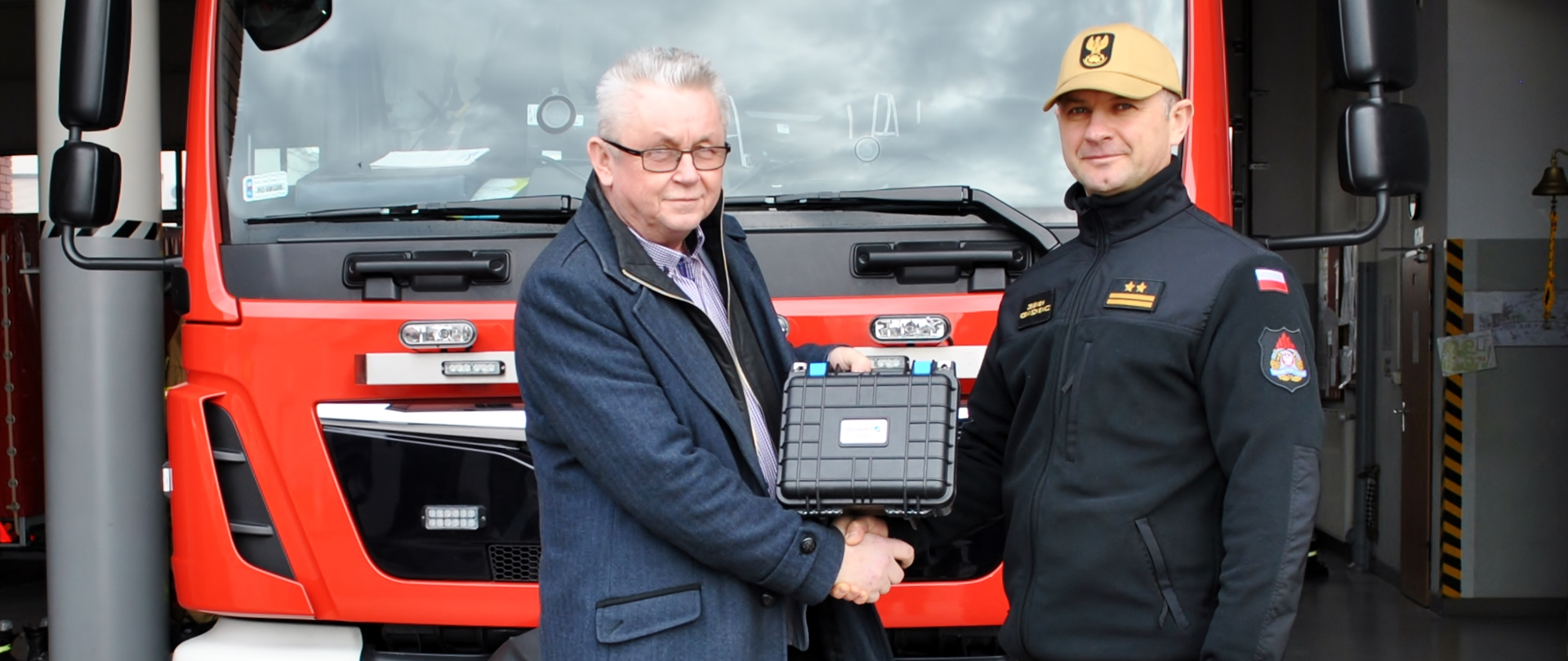 na tle samochodu stoją dwie osoby strażak i inna osoba w reku trzyma walizkę koloru czarnego 