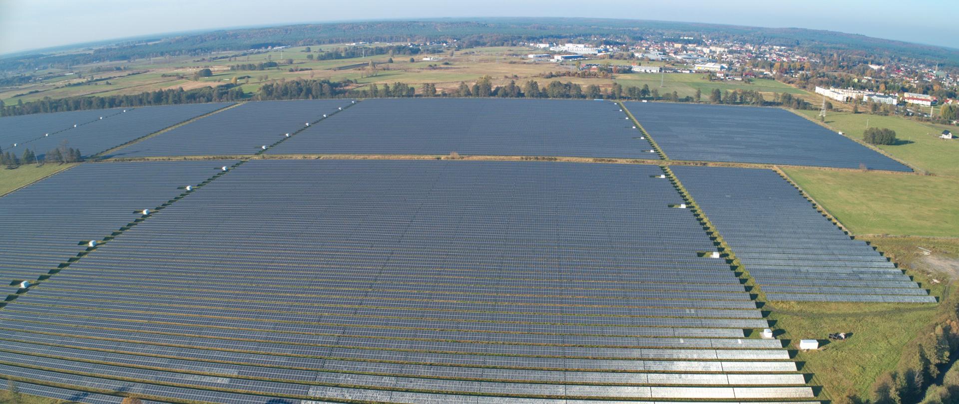 Krajobraz przedstawiający panele fotowoltaiczne na bardzo dużej powierzchni. W oddali drzewa i miejskie zabudowania.