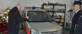 Przekazanie samochodu w OSP Jutrosin. Garaż w remizie OSP Jutrosin. proboszcz święci nowy nabytek strażaków - lekki samochód operacyjny marki citroen berlingo. W tle regał ze sprzętem, z prawej strony dwóch druhów OSP.