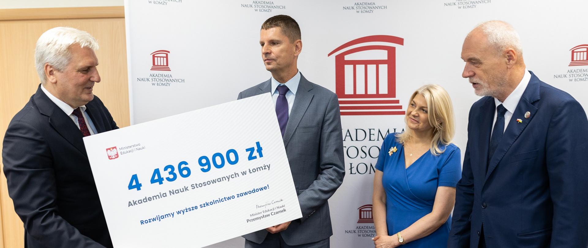 Minister Piontkowski stoi obok kobiety w niebieskiej sukni i dwóch mężczyzn w garniturach, wręcza jednemu z nich symboliczny czek z napisem 4 436 900 zł.
