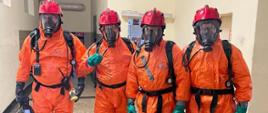 Wspólne zdjęcie policjantów ubranych w ubranie ochronne koloru pomarańczowego, aparaty ochrony układu oddechowego, kaski lekkie oraz rękawice ochronne. Policjanci stoją na korytarzu budynku.