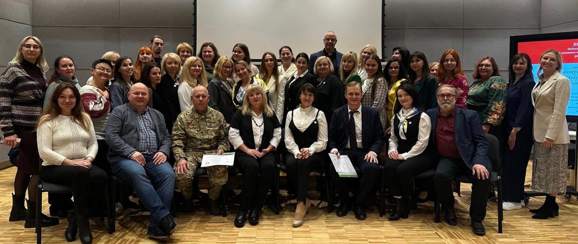 Ustawiona w trzy rzędy grupa kobiet i mężczyzn pozuje do zdjęcia na tle ściany z pustym ekranem