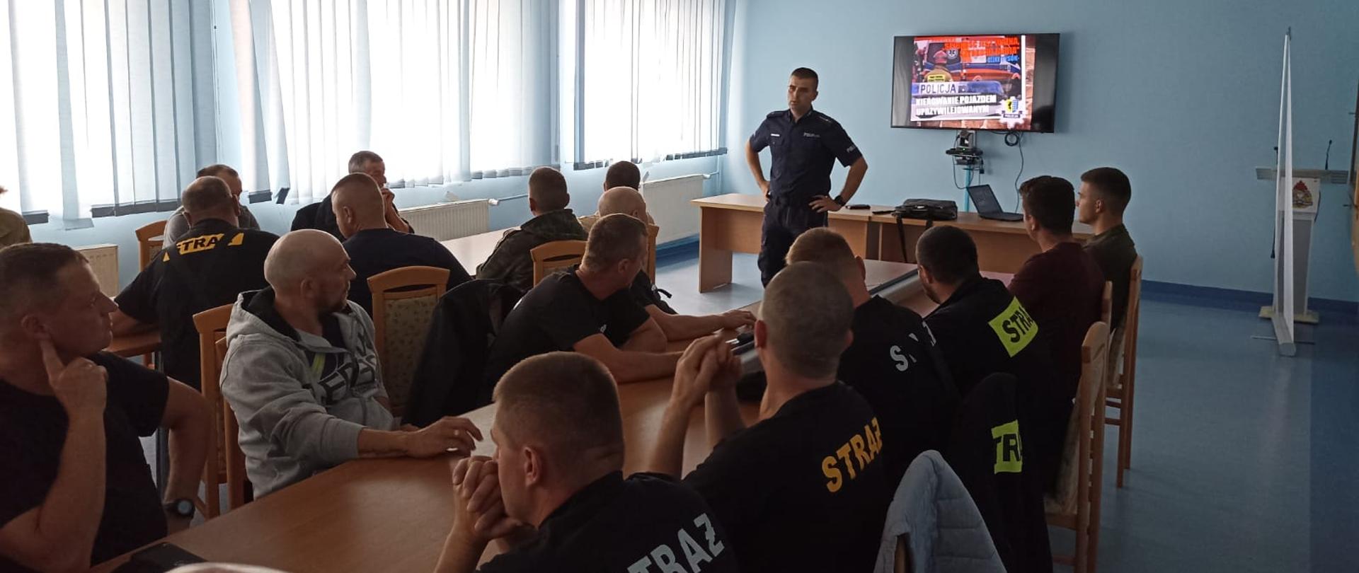 Kadr przedstawia szkolenie. Przy dwóch długich stołach siedzą strażacy. Patrzą się na prowadzącego. Prowadzącym spotkanie jest funkcjonariusz Policji. Za nim na telewizorze wyświetlana jest prezentacja. Pomieszczenie, w którym przebywają ma niebieskie ściany i podłogę. Z lewej strony okna zasłonięte są niebieskimi żaluzjami