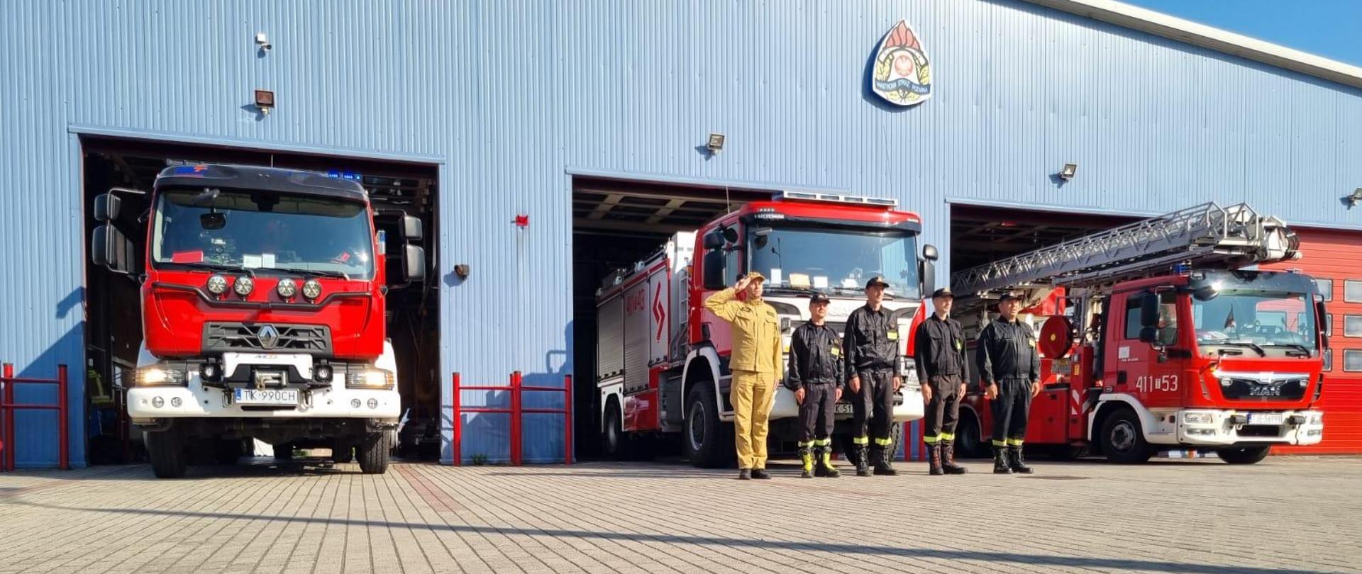 Zdjęcie obrazuje strażaków w umundurowaniu koszarowym, którzy przed garażem jednostki ratowniczo-gaśniczej oddają hołd bohaterom Powstania Warszawskiego. W otwartych bramach garażowych widać samochody ratowniczo-gaśnicze z włączonymi sygnałami świetlnymi. Jeden ze strażaków oddaje honor.