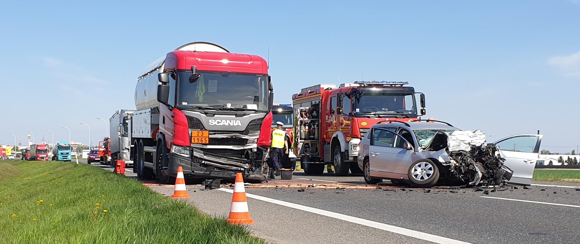 Na zdjęciu widać dwa rozbite pojazdy: ciężarowy pojazd, autocysterna z czerwona kabiną oraz srebrny pojazd osobowy. W pobliżu widać pojazdy straży pożarnej w kolorze czerwonym, oznakowane biało z sygnalizacją ostrzegawczą w kolorze niebieskim. Niebo błękitne. Na asfalcie porozrzucane fragment karoserii. Rozsypany sorbent w kolorze różowym. 