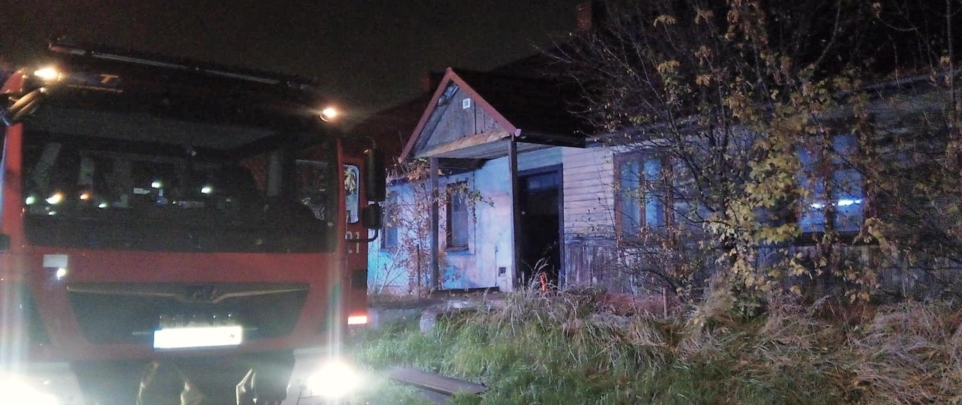 Na zdjęciu po lewej stronie stoi samochód pożarniczy a po prawej widać opuszczony dom jednorodzinny, w którym doszło do pożaru drzwi wejściowych.