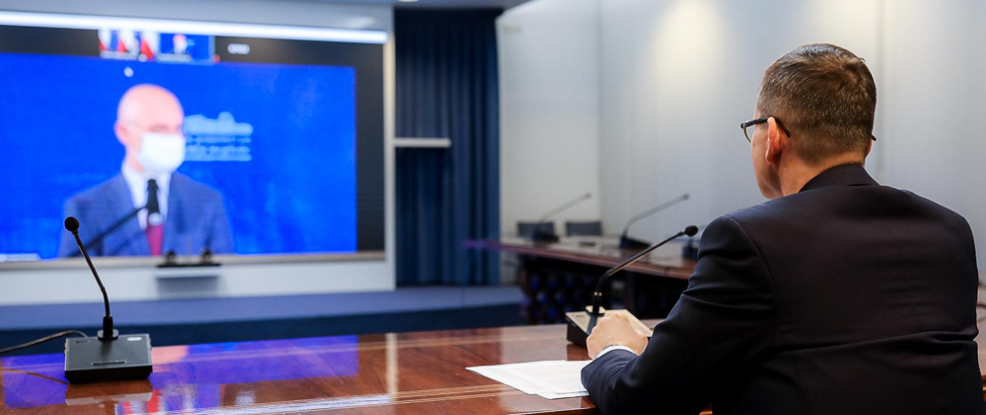 Na pierwszym planie premier Mateusz Morawiecki siedzi przy stole, na którym ustawiony jest mikrofon. W tle na ekranie widać ministra Michała Kurtykę.