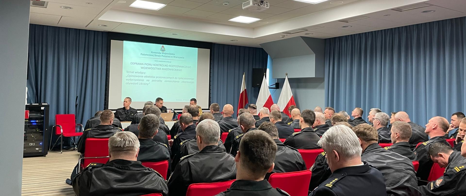 odprawa pionu kontrolno-rozpoznawczego PSP województwa mazowieckiego. uczestnicy siedzą na krzesłach w sali konferencyjnej. na przodzie przy mównicy siedzi zastępca mazowieckiego komendanta PSP oraz naczelnicy wydziału kontrolno-rozpoznawczego komendy wojewódzkiej psp w warszawie. w tle wyświetlana jest prezentacja na rzutniku.