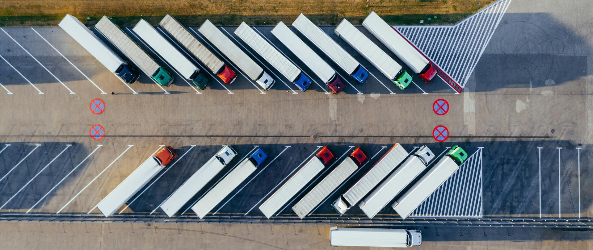 Na dużym parkinku przy autostradzie stoją w dwóch rzędach zaparkowane TIRy. Zdjęcie zrobione jest z lotu ptaka
