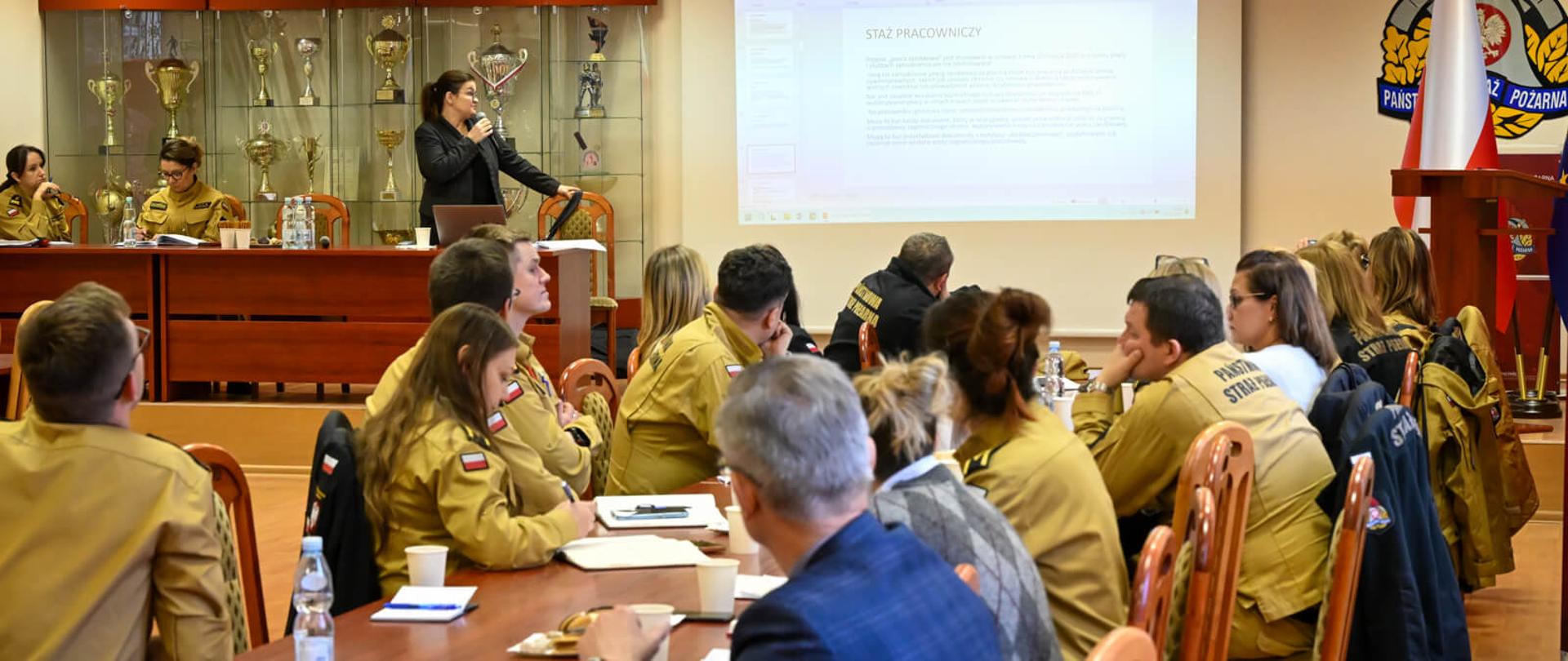 ujęcie z sali szkoleniowej. Kobieta w ciemnym stroju przemawia do grupy słuchaczy siedzących przy stole, podczas gdy na dużym ekranie wyświetlana jest prezentacja z nagłówkiem "Staż pracowniczy". W tle widać gablotę z pucharami.