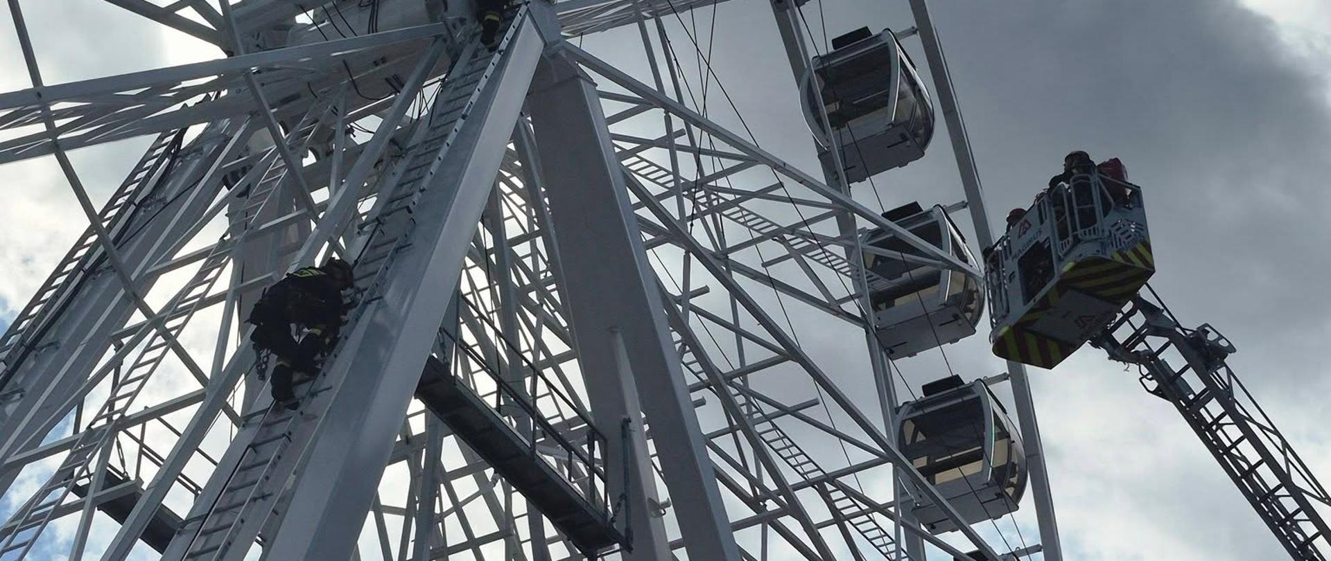 Strażacy wykonują ćwiczenia wysokościowe na konstrukcji koła widokowego. Jeden z ratowników wspina się po drabinie, podczas gdy drugi znajduje się poniżej i zabezpiecza jego działania. Wokół widoczna jest stalowa konstrukcja urządzenia.
