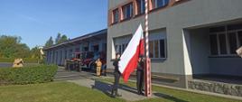 Na zdjęciu na tle budynku Komendy Miejskiej Państwowej Straży Pożarnej w Częstochowie i Jednostki Ratowniczo-Gaśniczej nr 2 maszt flagowy. Przy maszcie stoi poczet flagowy podnoszący flagę. W oddali widać ustawionych strażaków i wozy bojowe.