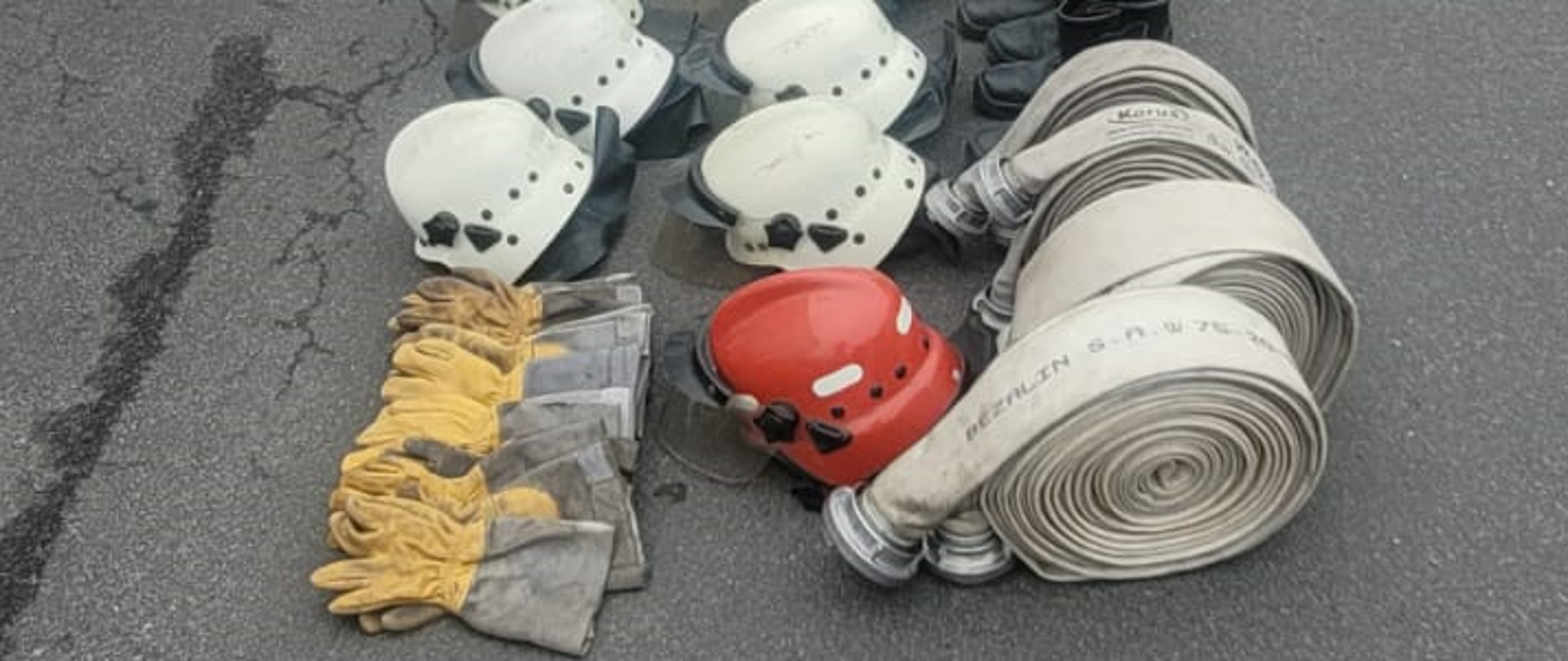 Zdjęcie przedstawia rękawice, hełmy buty i węże pożarnicze leżące na ziemi, hełmy koloru białego i czerwonego