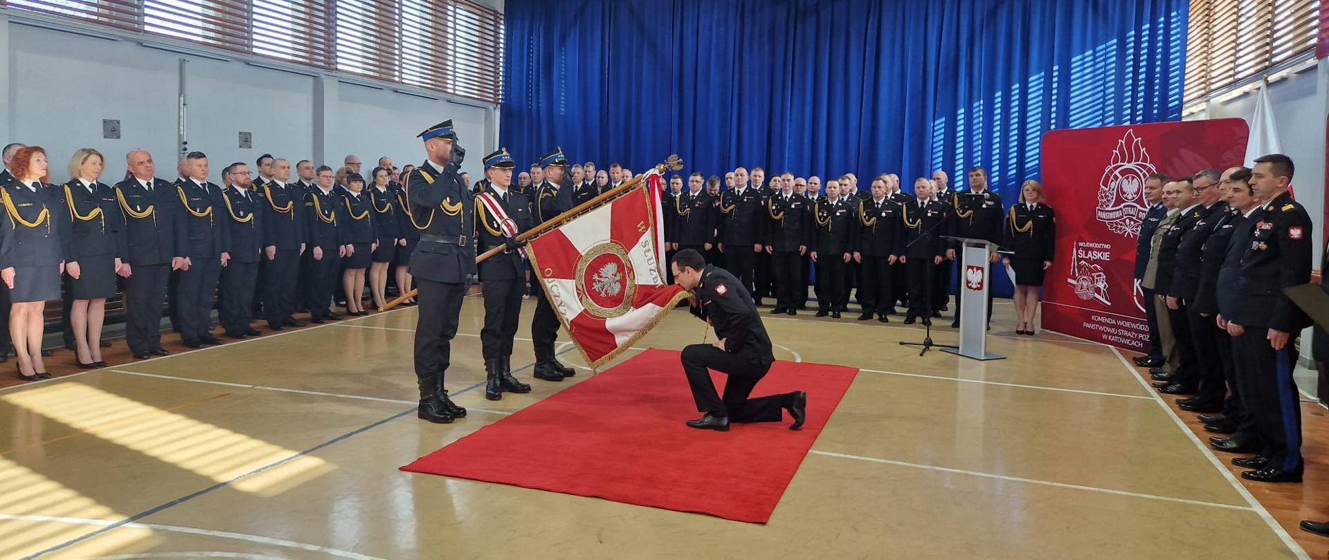 Moment powitania nowego śląskiego komendanta wojewódzkiego ze sztandarem. Na sali gimnastycznej widoczni zgromadzeni funkcjonariusze i funkcjonariuszki w umundurowaniu galowym oraz zaproszeni goście, mężczyźni w garniturach i mundurach innych formacji