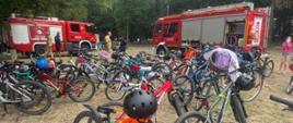 Rowery dzieci które uczestniczyły w kolonii i przybyły z policjantami nad wodę. Na drugim planie samochody bojowe PSP w otoczeniu parku.