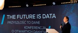 Wiceminister Adam Andruszkiewicz przemawia z pulpitu. W tle napis: The future is data. Przyszłość to dane. Konferencja o otwartych danych.