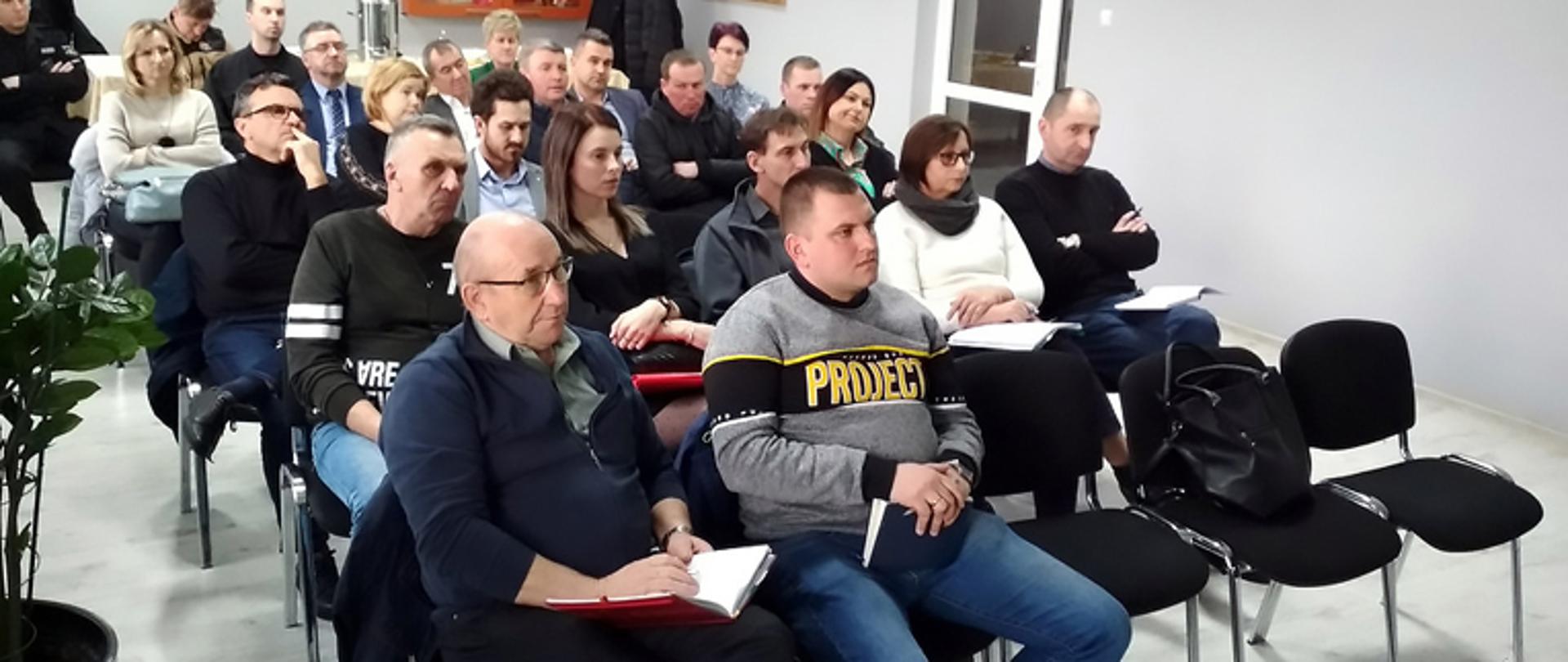 Wójtowie oraz prezesi Zarządów Gminnych ZOSP RP podczas spotkania z komendantem powiatowym PSP w Łowiczu, na sali siedzą w kilku rzędach kobiety i mężczyźni, w tle gablota ze sztandarek komendy powiatowej PSP w Łowiczu i stół na którym stoi duży metalowy termos, po lewej stronie wieszak na odzież, na pierwszym planie po lewej stronie stoi na podłodze duży kwiat w dużej szarej donicy