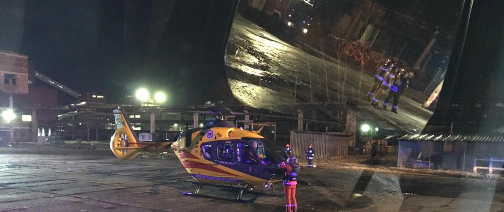 Zdjęcie przedstawia Helikopter LPR na placu wewnętrznym kopalni przygotowujący się do startu i strażaków zabezpieczających start helikoptera.