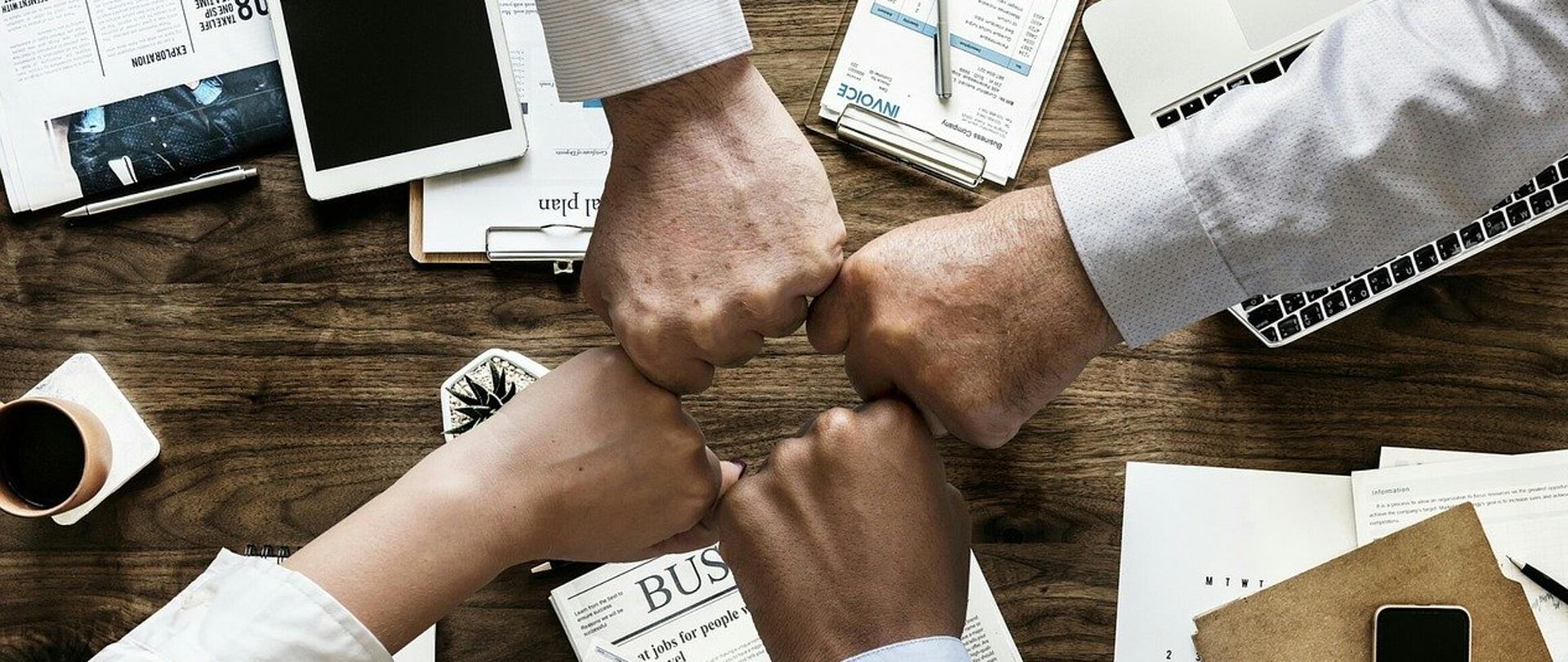 Cztery osoby, w tym jedna kobieta, stykają się pięściami nad centralną częścią drewnianego stołu. Na stole leży otwarty laptop, tablet i telefon komórkowy. Wśród nich znajdują równomiernie porozrzucane przedmioty biurowe: kalkulator, białe, niezapisane kartki, dokumenty z zestawieniami tabelarycznymi, gazety, długopisy, kubek z kawą oraz roślina w doniczce.