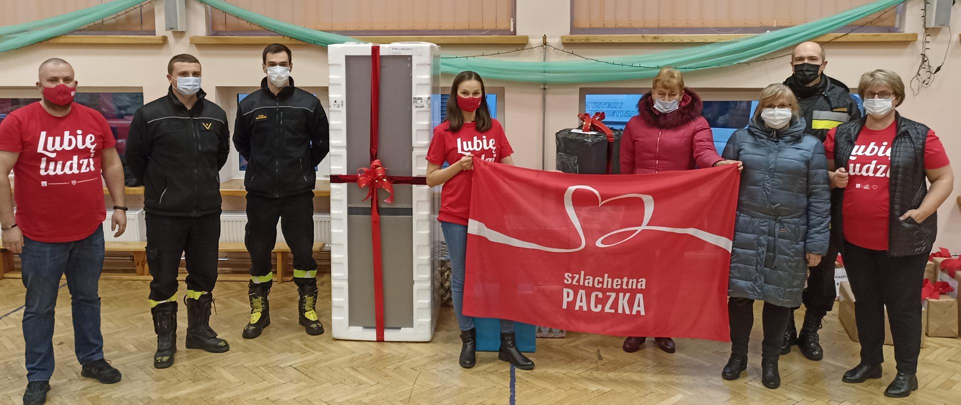 Na zdjęciu widzimy pracowników oraz funkcjonariuszy PSP, którzy przekazują organizatorom akcji szlachetna paczka zakupione dary. 