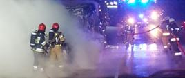Zdjęcie wykonane w porze nocnej. Fotografia przedstawia dwa samochody (osobowy i bus) stojące przodami do siebie (po wypadku) na jezdni w trakcie pożaru. Strażacy (5 osób) przy użyciu dwóch linii gaśniczych prowadzą działania gaśnicze. Za osobami widoczny samochód strażacki w włączoną sygnalizacją świetlną (niebieską).