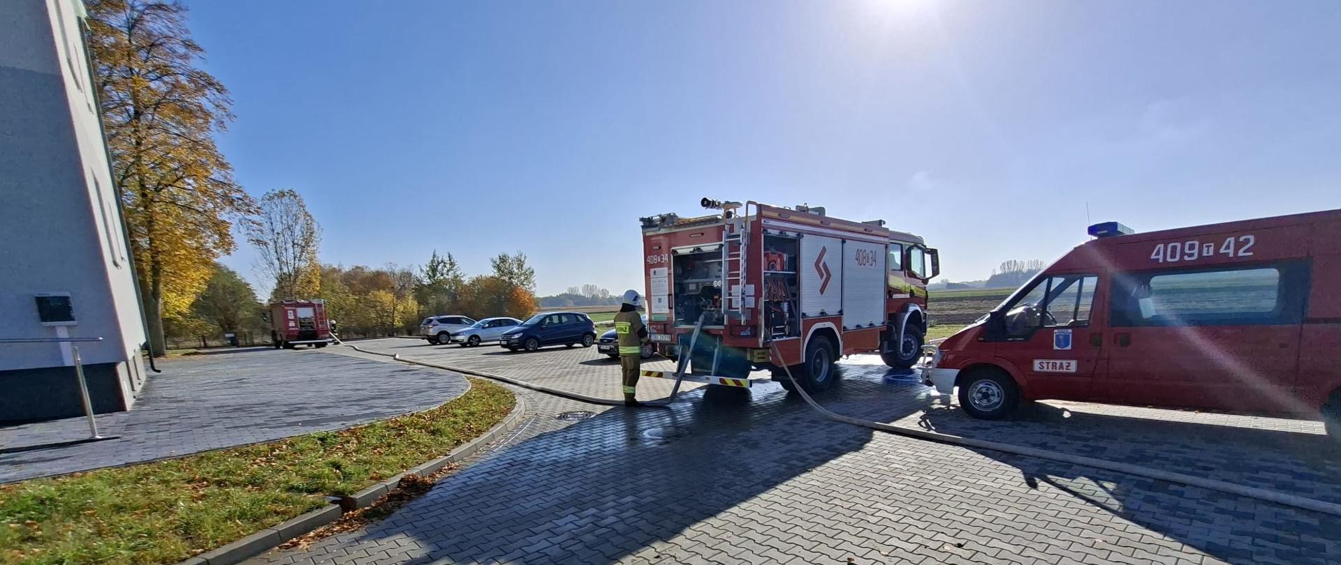 Widok z zewnątrz na plac manewrowy lub parking przed budynkiem w słoneczny dzień. Na miejscu pracują dwa czerwone pojazdy straży pożarnej – wóz bojowy oraz mniejszy samochód operacyjny z oznaczeniami straży (np. numerem bocznym 409[wężyk]42). Od wozu pożarniczego ciągną się węże strażackie, a w tle widać zaparkowane samochody osobowe oraz drzewa jesienią.
