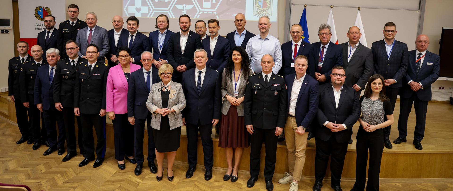 Zdjęcie grupowe uczestników Wyższego Kursu Ochrony Ludności w Akademii Pożarniczej, na tle ekranu z nazwą kursu i flag narodowych.