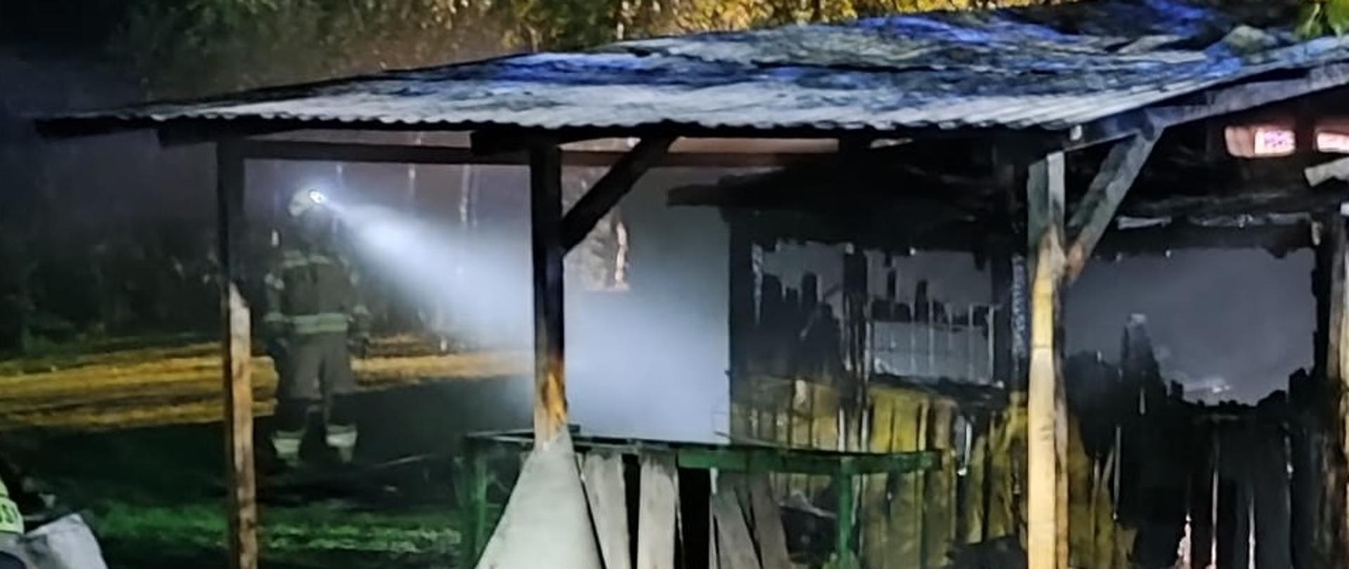 Spalony domek letniskowy. Przed domkiem widoczne są dwa odcinki wężowe a zanim stoi strażak i sprawdza pogorzelisko. 