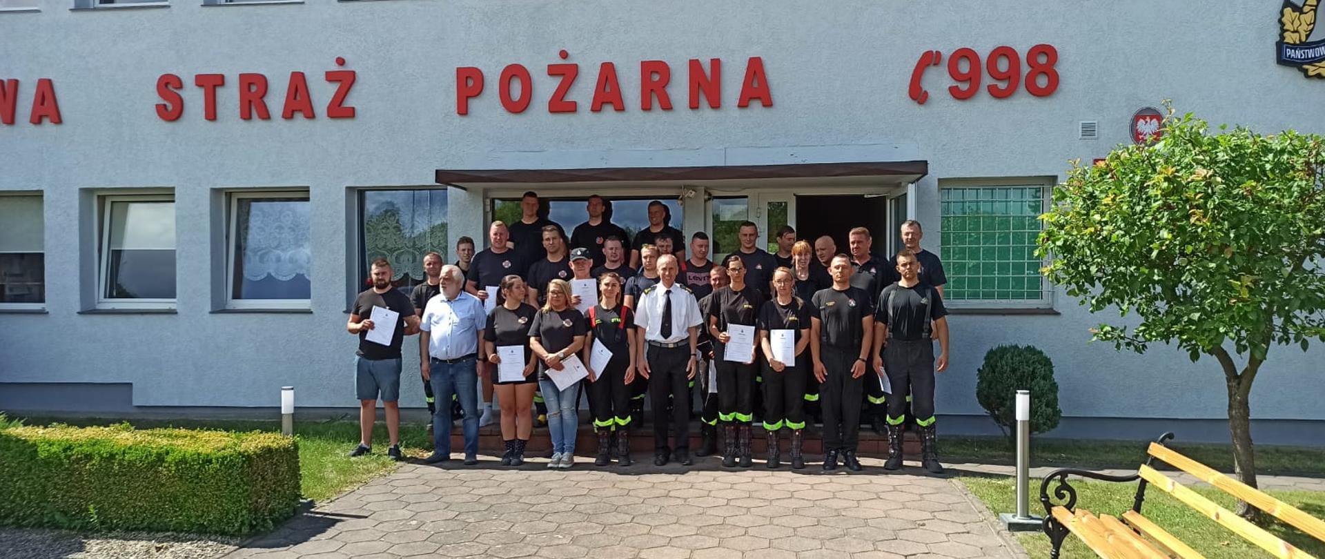 Zdjęcie przedstawia grupę ludzi przed budynkiem komendy straży pożarnej w Świdwinie. Na ścianie widzimy napis Straż Pożarna 998.